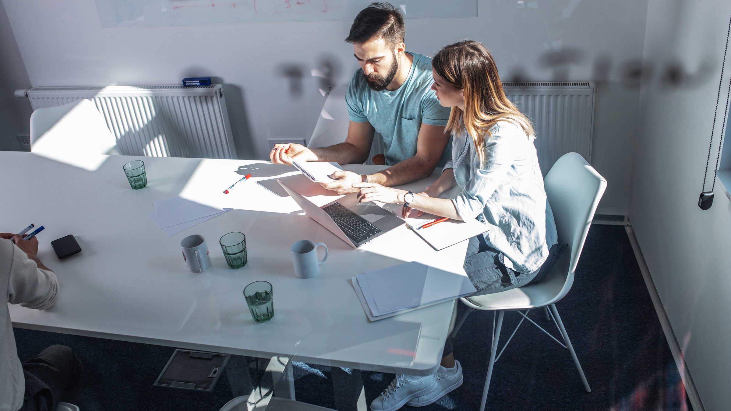 Two colleagues at a laptop on a white table, a whiteboard behind them