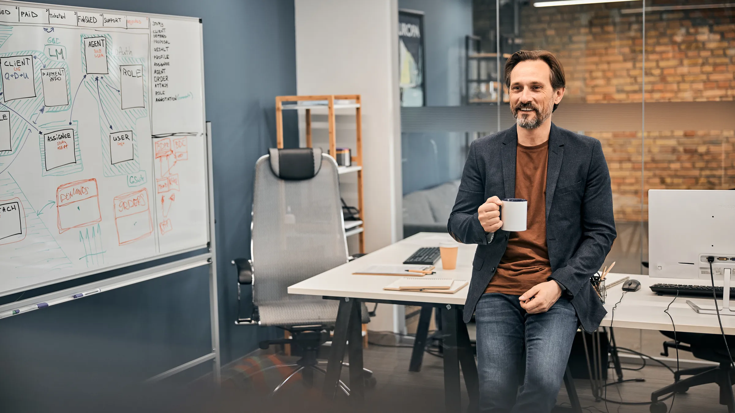 A smiling leader with a coffee mug leans on a desk beside a whiteboard of system sketches