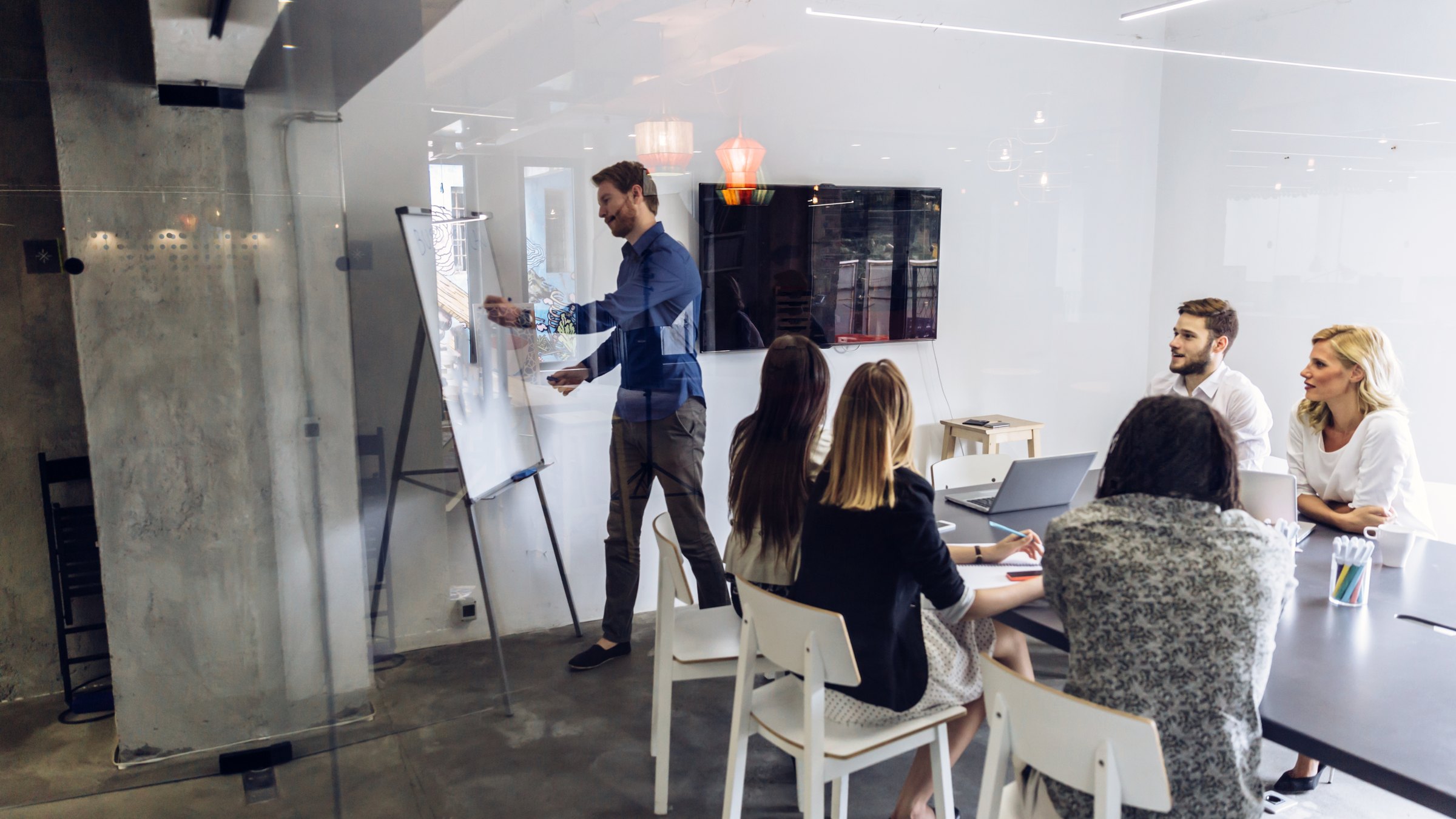 A presenter stands at a flip chart while colleagues sit around a table in a glass-walled meeting room