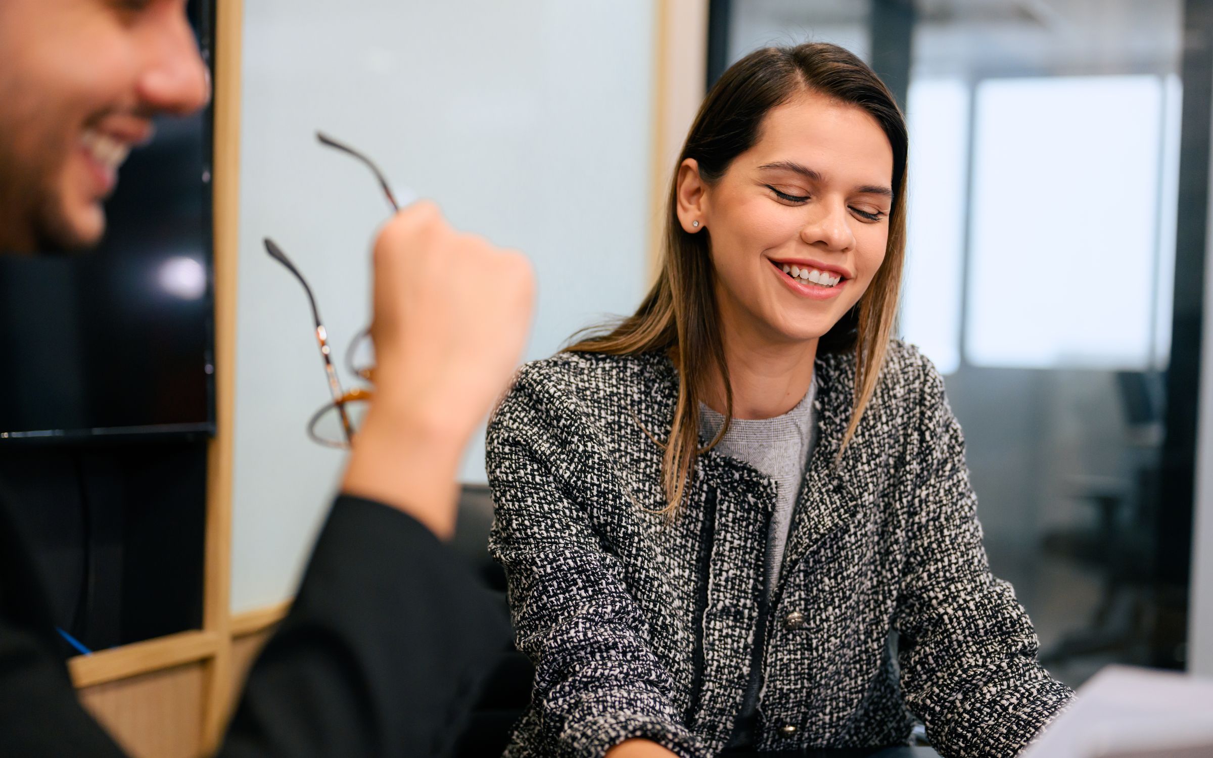 A woman in a tweed jacket smiling in an office, with a colleague holding glasses in the foreground.
