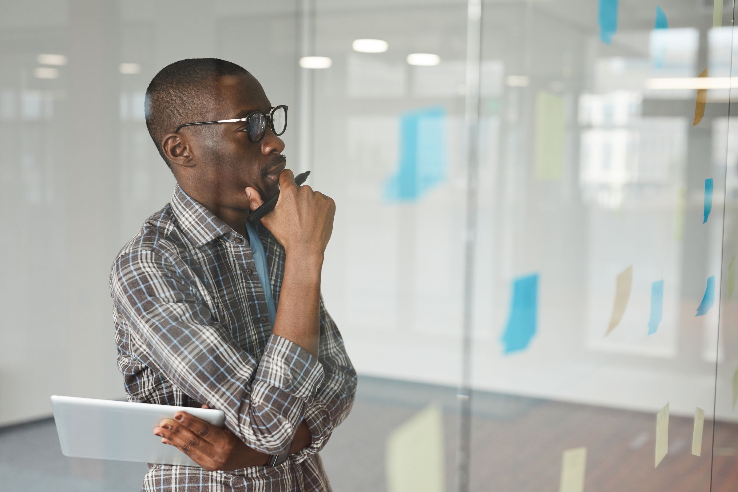 A man in glasses and a plaid shirt holding a tablet, looking at sticky notes on a glass wall.