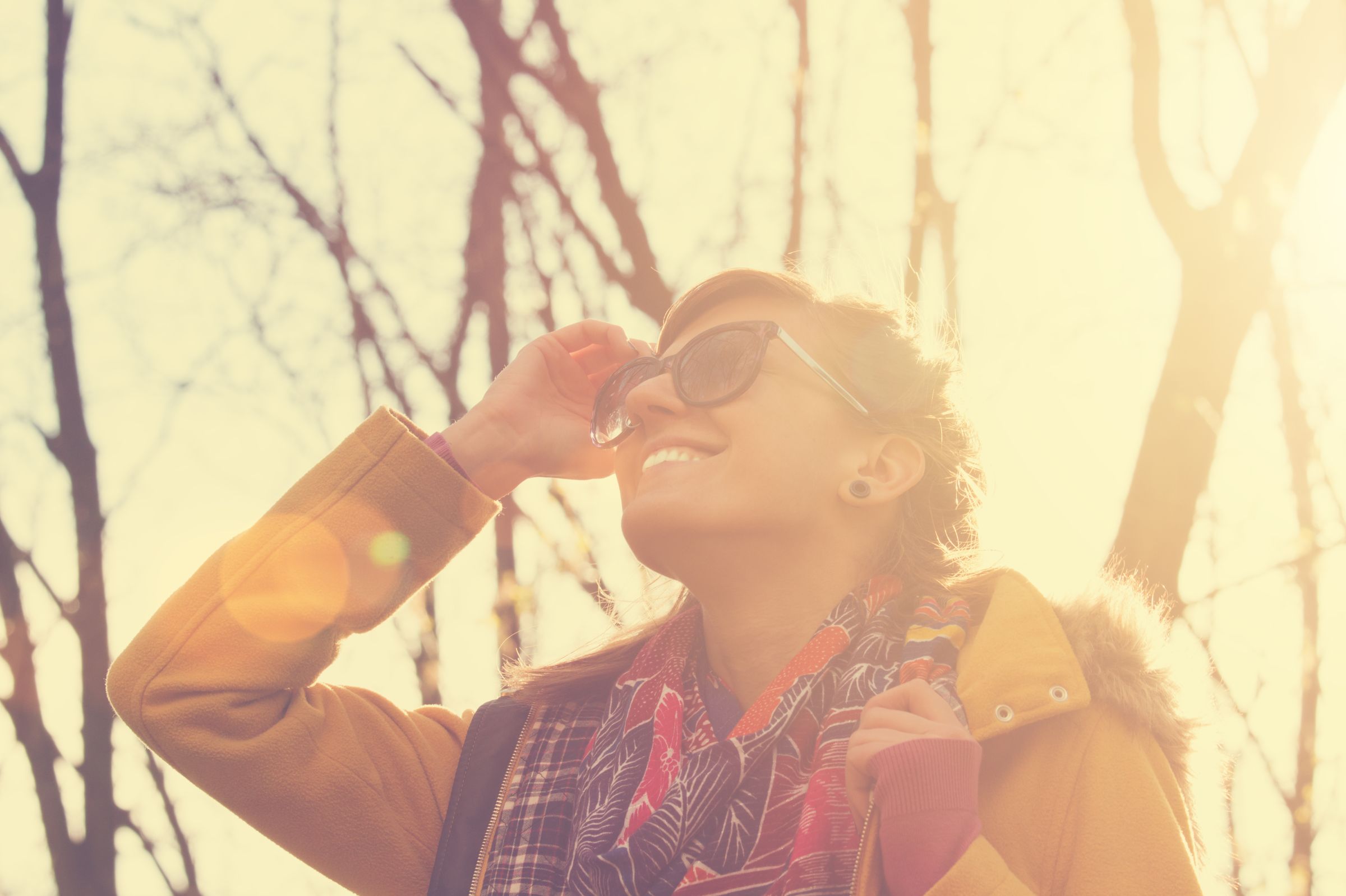 A woman in sunglasses smiling against backlit trees.