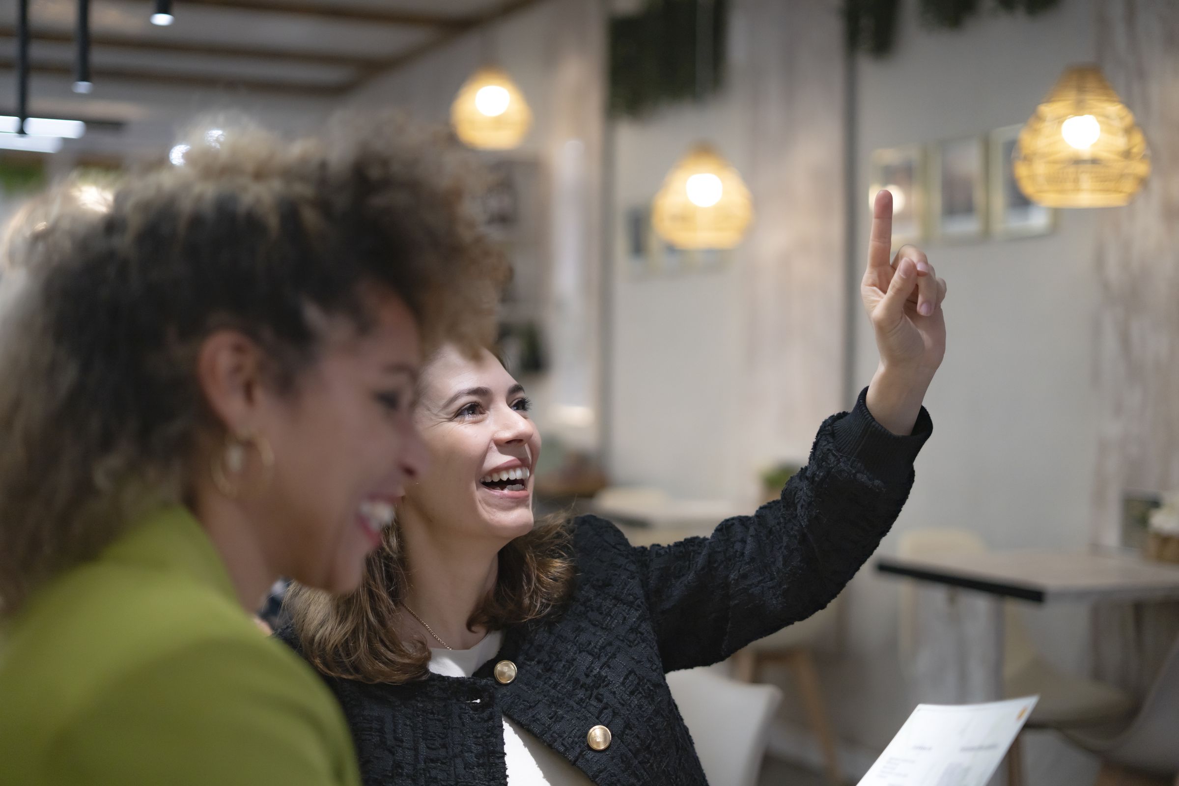 Two women laughing in a cafe, one in a dark jacket pointing up.
